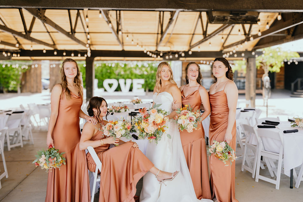 Bride surrounded by five bridesmaids in satin copper dresses, all holding pastel floral bouquets under a wooden pergola at The Venue at Chilleens.