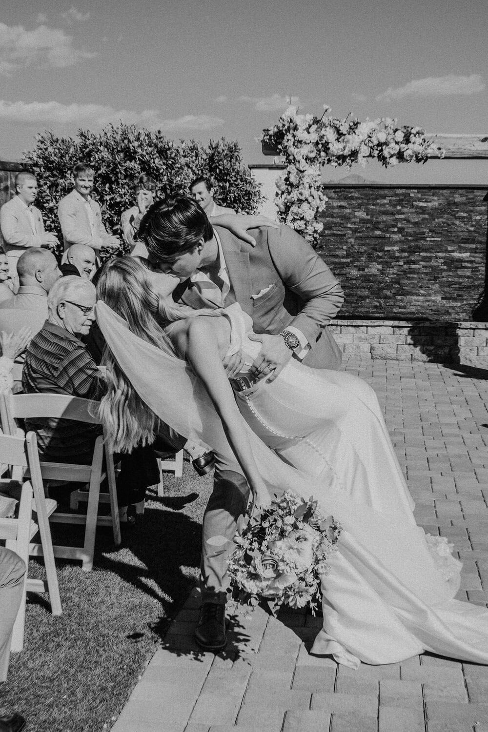 A Swoon-Worthy Sendoff Groom dipping bride into a romantic kiss as guests look on, captured in black and white at the end of the ceremony aisle.
