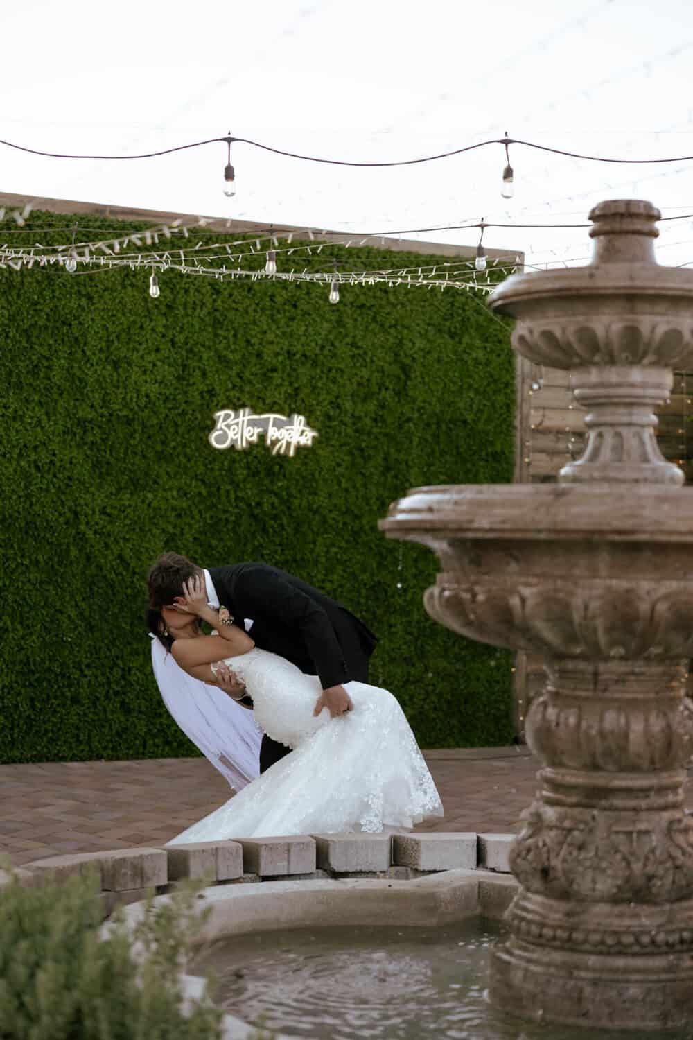 Bride and groom kissing in front of lush green wall at The Venue at Chilleens, holding white rose bouquet, Arizona wedding portrait.