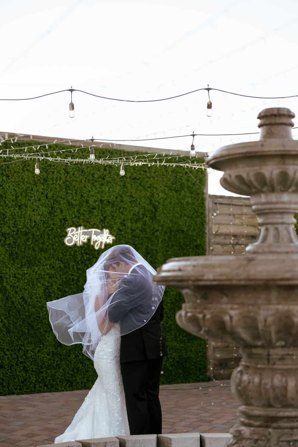 Bride and groom kissing in front of lush green wall at The Venue at Chilleens, holding white rose bouquet, Arizona wedding portrait. with veil, at the Venue at Chilleens